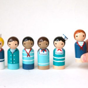 5 wooden sailor toy figurines and 1 being held by a child’s hand. 1 pale skinned boy with blond curly hair and a hat, 1 pale skinned boy with brown hair, 1 medium skinned boy with black hair, 1 medium skinned girl with brown pigtails, 1 pale skinned girl with black hair, 1 pale skinned girl with red hair.