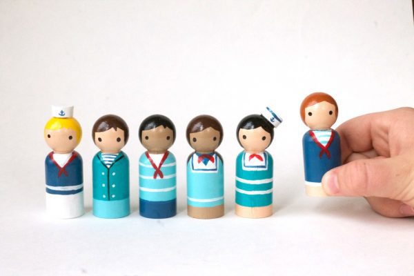5 wooden sailor toy figurines and 1 being held by a child’s hand. 1 pale skinned boy with blond curly hair and a hat, 1 pale skinned boy with brown hair, 1 medium skinned boy with black hair, 1 medium skinned girl with brown pigtails, 1 pale skinned girl with black hair, 1 pale skinned girl with red hair.