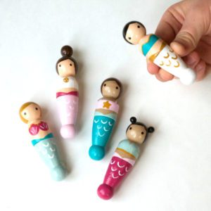 4 wooden mermaid peg dolls laying down and 1 being held by a child’s hand. 1 pale skinned with blond hair, 1 pale skinned with a brown top knot, 1 medium skinned with brown pigtails, 1 medium skinned with black double buns.