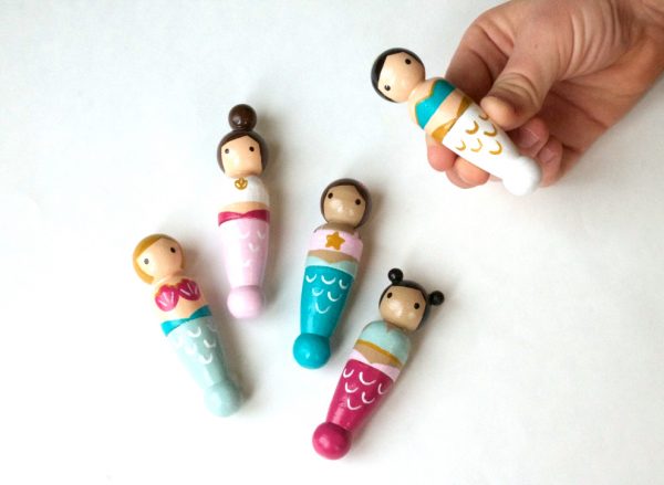 4 wooden mermaid peg dolls laying down and 1 being held by a child’s hand. 1 pale skinned with blond hair, 1 pale skinned with a brown top knot, 1 medium skinned with brown pigtails, 1 medium skinned with black double buns.