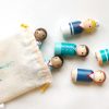 6 nautical peg dolls lay down, coming out of a bag. 1 pale skinned boy with blond curly hair and a hat, 1 pale skinned boy with brown hair, 1 medium skinned boy with black hair, 1 medium skinned girl with brown pigtails, 1 pale skinned girl with black hair, 1 pale skinned girl with red hair.