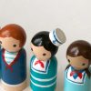 close up of 6 nautical peg dolls standing in a row. 1 pale skinned boy with blond curly hair and a hat, 1 pale skinned boy with brown hair, 1 medium skinned boy with black hair, 1 medium skinned girl with brown pigtails, 1 pale skinned girl with black hair, 1 pale skinned girl with red hair.