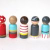 back view of 5 wooden pirate peg dolls standing in a row. 1 pale skinned with pirate hat, 1 medium skinned with bandana, 1 pale skinned with eye patch and bandana, 1 pale skinned with head wrap, 1 medium skinned who is bald with a dark beard