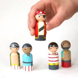 4 wooden pirate peg dolls standing in a row, and one being held by a child's hand. 1 pale skinned with pirate hat, 1 medium skinned with bandana, 1 pale skinned with eye patch and bandana, 1 pale skinned with head wrap, 1 medium skinned who is bald with a dark beard