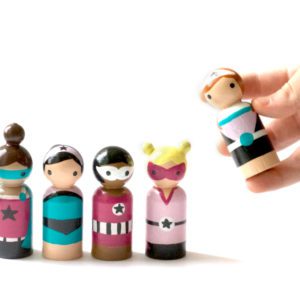 5 superhero girl peg dolls standing in a row. One is being picked up by a child. 1 doll with dark skin and brown hair, 1 doll with pale skin and black hair, 1 doll with dark skin and black hair, 1 doll with pale skin and blond hair, 1 doll with pale skin and red hair.
