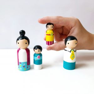 3 Asian family peg dolls standing in a row and 1 being held by a child’s hand. 1 mother, 1 boy, 1 girl, and 1 dad