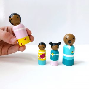 3 dark skinned family dolls standing in a row and one being held by a child’s hand. 1 mother, 1 boy, 1 girl, and 1 dad