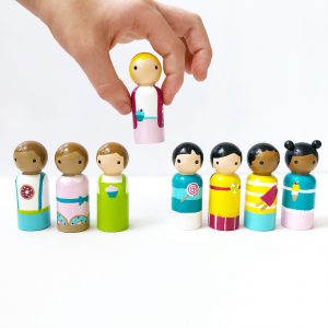 Multicultural peg dolls set with 7 dolls standing in a row and 1 being held by a child’s hand. 1 boy with medium skin and brown hair, 1 girl with medium skin and brown hair, 1 boy with pale skin and brown hair, 1 girl with pale skin and blond hair, 1 boy with pale skinned and black hair, 1 girl with pale skinned and black pig tails, 1 boy with dark skin and black hair, 1 girl with dark skinned and black hair buns.