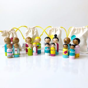 4 multicultural peg people families standing in a row in front of bags. Each family has a mom, a dad, a girl, and a boy. 1 medium skinned family with brown hair, 1 pale skinned family with brown and blond hair, 1 pale skinned family with black hair, 1 dark skinned family with black hair.