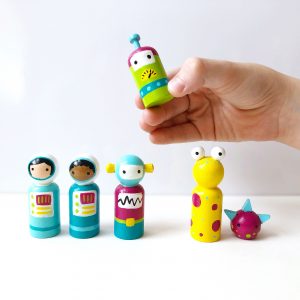 5 wooden space toys for kids standing and 1 being held by a child’s hand. 1 pale skinned female astronaut, 1 dark sinned male astronaut, 2 robots, and 2 aliens