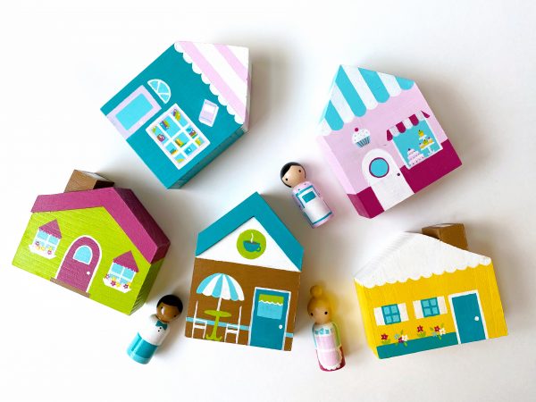 5 wooden toy block houses and 3 peg dolls laying down. 1 bookstore with worker, 1 cafe with worker, 1 bakery with worker, 1 yellow house and 1 green house.