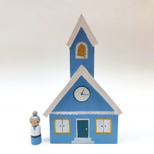 blue mini wooden schoolhouse toy with pale skinned, gray haired teacher standing next to it.